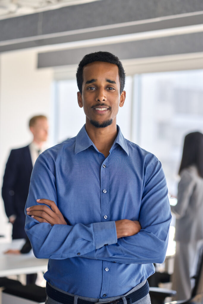 Confident young African American business man worker standing in office looking at camera. Smiling businessman employee corporate leader executive in meeting room with team, vertical portrait.